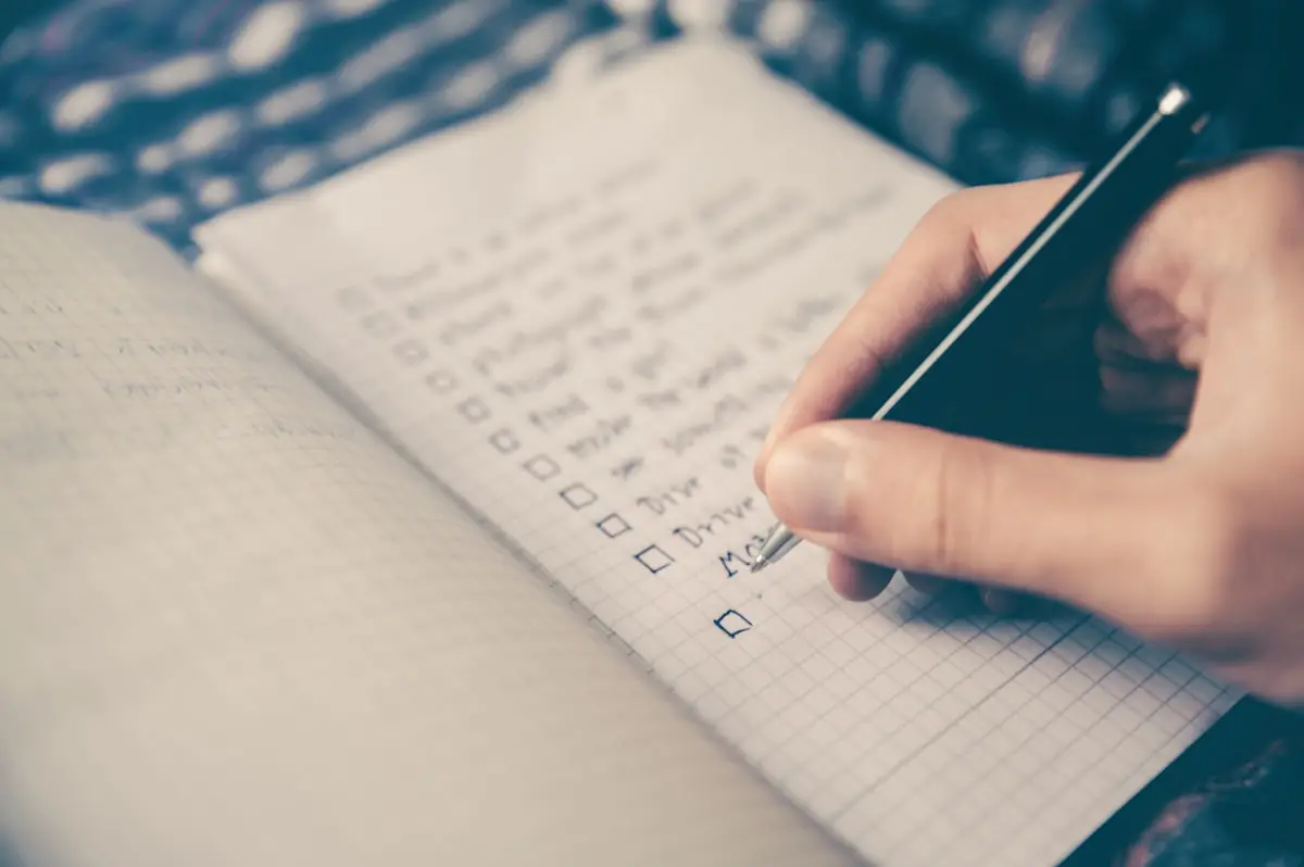 The Krehzy Good Guide to Resetting Your Business: An End-of-Year Systems Checklist for Entrepreneurs 2 A close-up of a hand holding a pen, writing a checklist in a grid-lined notebook. Several items are listed, some with checkboxes ticked, on top of a patterned fabric background.