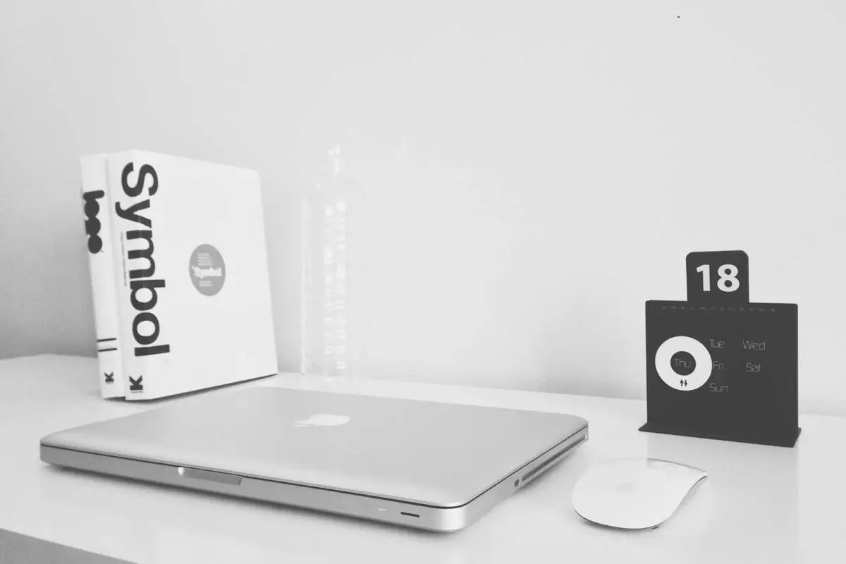 Why Church Volunteer Scheduling is Broken (And How to Fix It Without Hiring Staff) 2 A minimalistic white desk with a closed laptop, wireless mouse, books, a clear water bottle, and a small black calendar displaying the number 18.