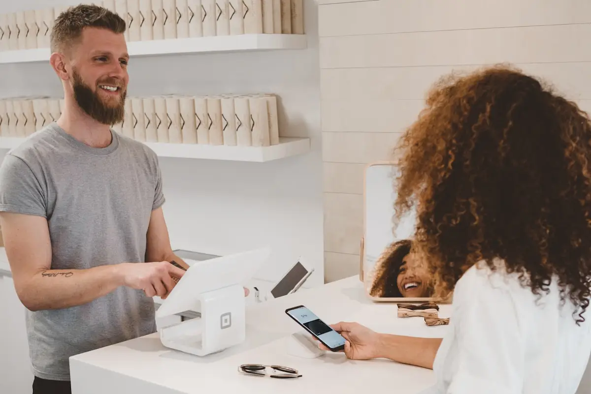 The Krehzy Good Guide to Client Automation: Why Enhancing Your Business Isn't Impersonal 2 A smiling cashier with a beard assists a customer making a contactless payment with a smartphone at a modern, minimalistic retail counter. Sunglasses and shopping bags are visible on the counter and shelves.