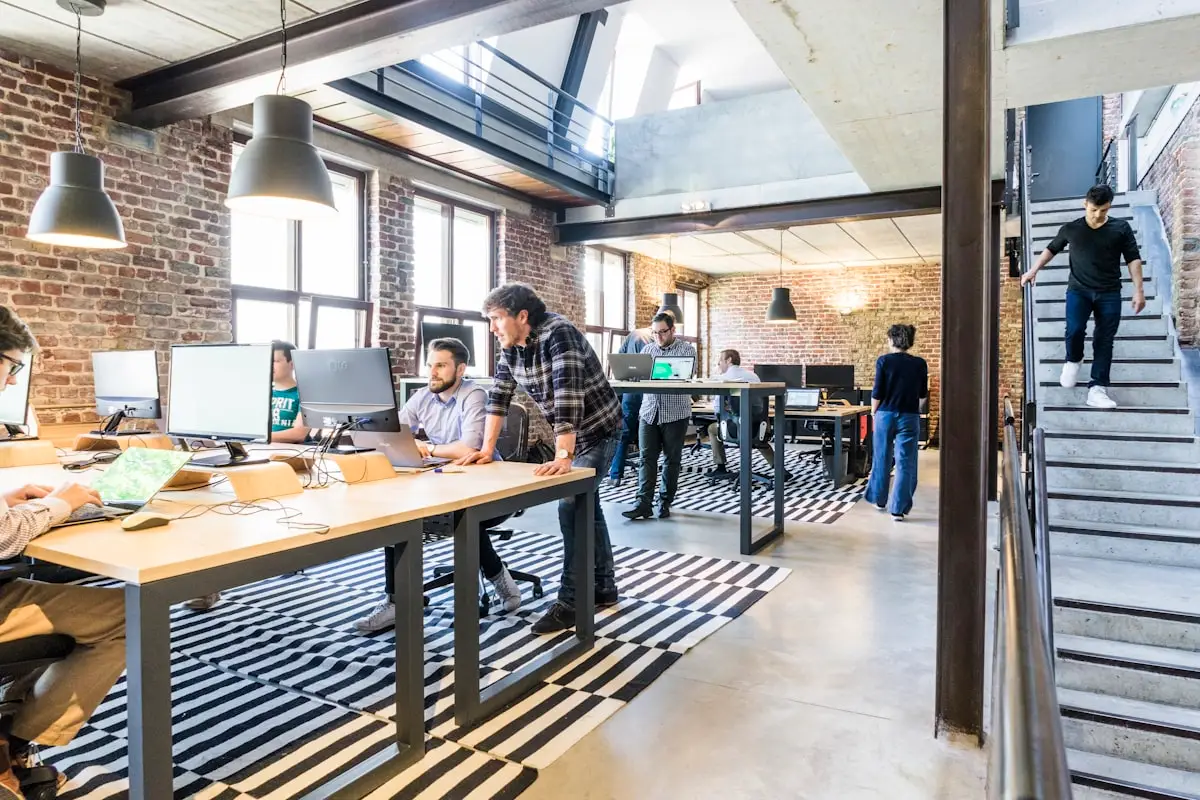 The Krehzy Good Guide to Reclaiming Your Time: Top 5 Automations for Solopreneurs 2 Open-plan office with people working at computers, having discussions, and walking. Exposed brick walls, large windows, modern furniture, and striped rugs create a bright, collaborative workspace atmosphere.