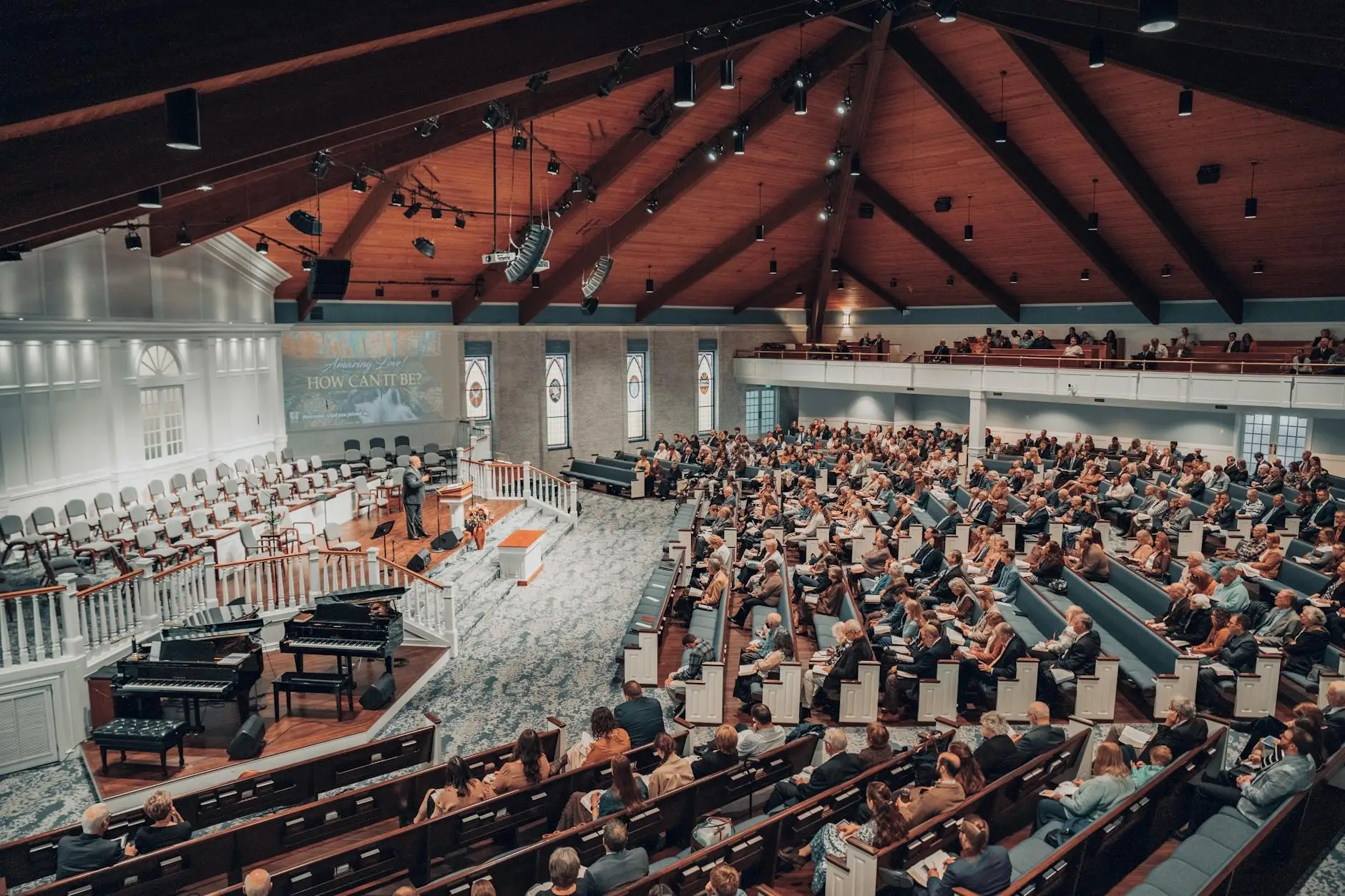 End the Staff Meeting Chaos 2 A large congregation sits in pews inside a spacious church with a high wooden ceiling, stage, and musical instruments. People face forward, listening attentively to a speaker or presentation at the front.